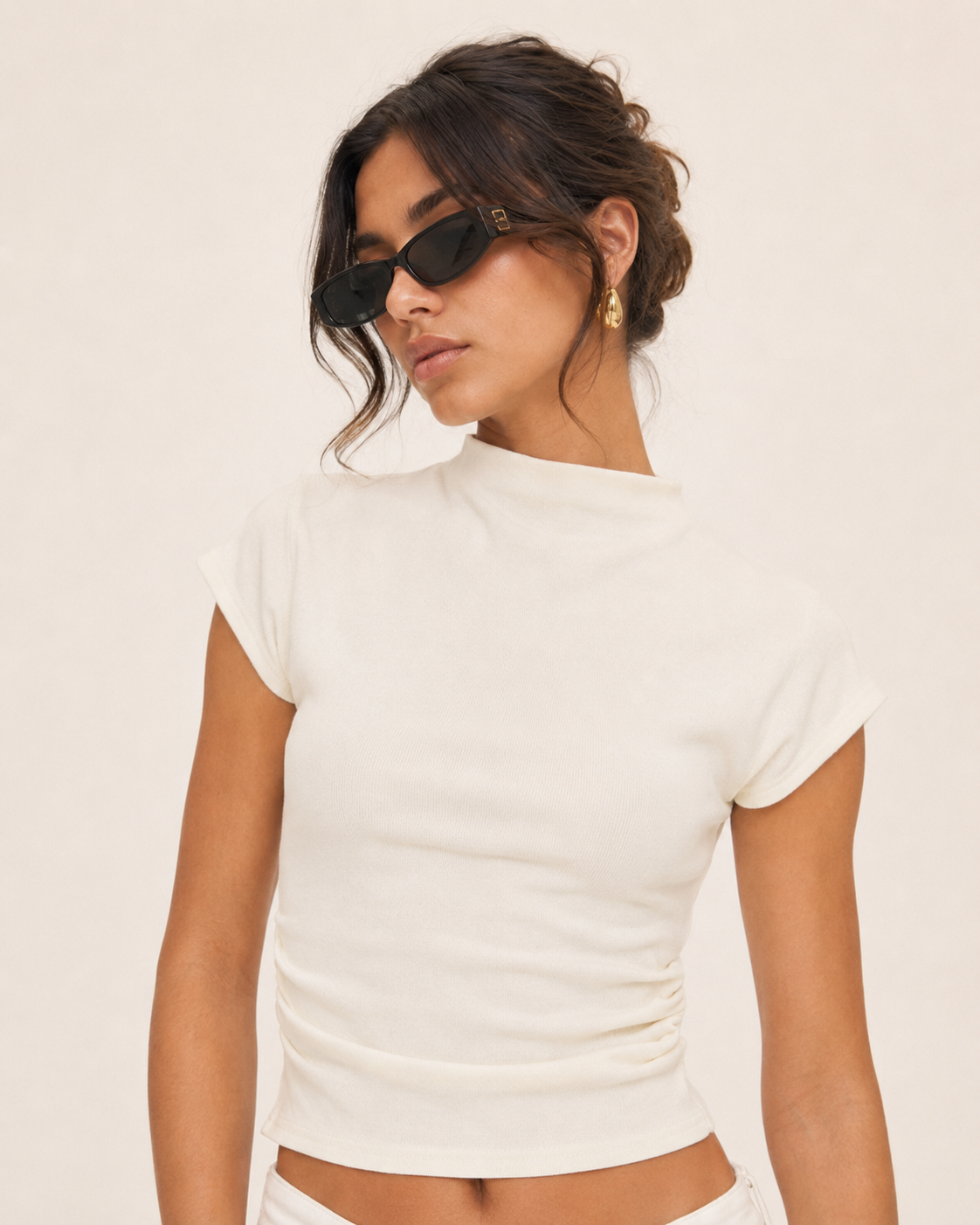 A woman wearing a fitted short-sleeve off-white ribbed crop top and black sunglasses, photographed against a plain light beige studio background.